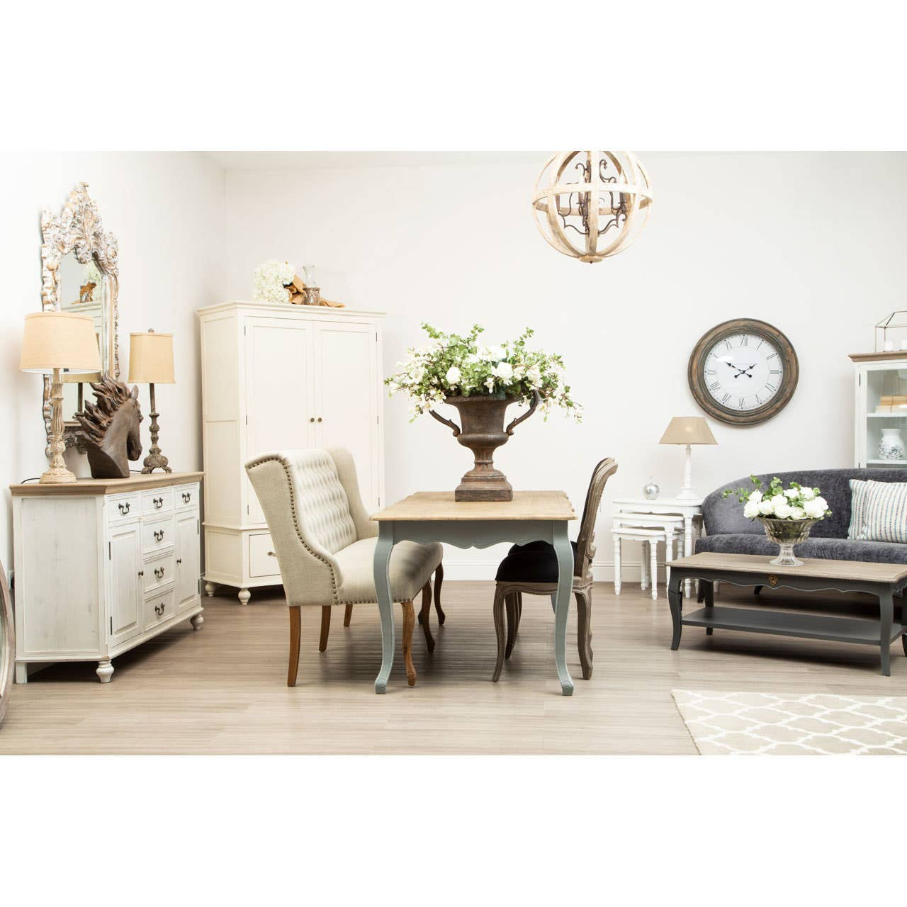 A stylish grey velvet sofa with three seats, featuring plush cushions and a sleek design, positioned in a bright, modern living space. The room includes a dining table with chairs, a decorative vase with flowers, a cabinet, and a wall clock, all set against a light-colored backdrop