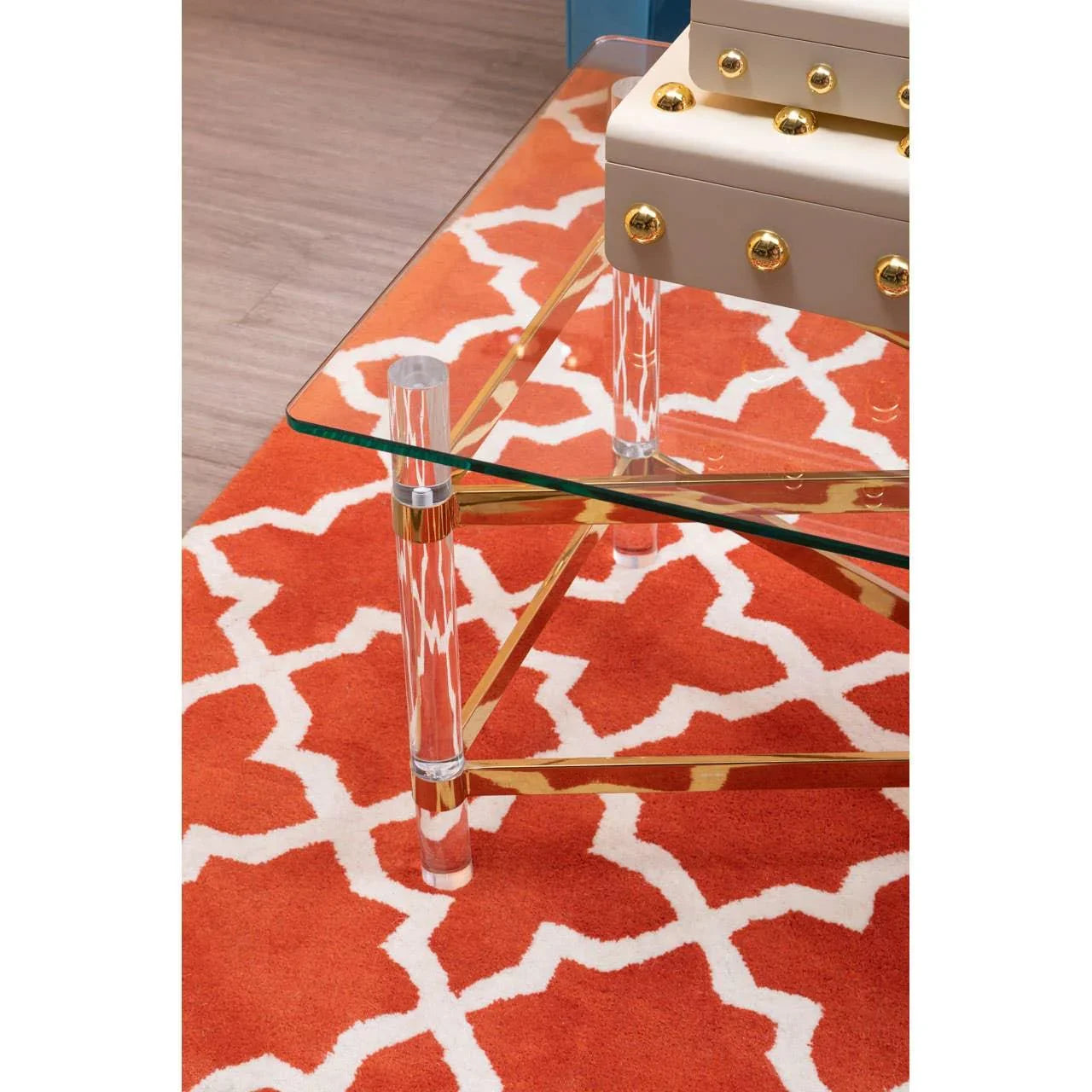Glass coffee table with gold trim and acrylic legs on orange patterned rug
