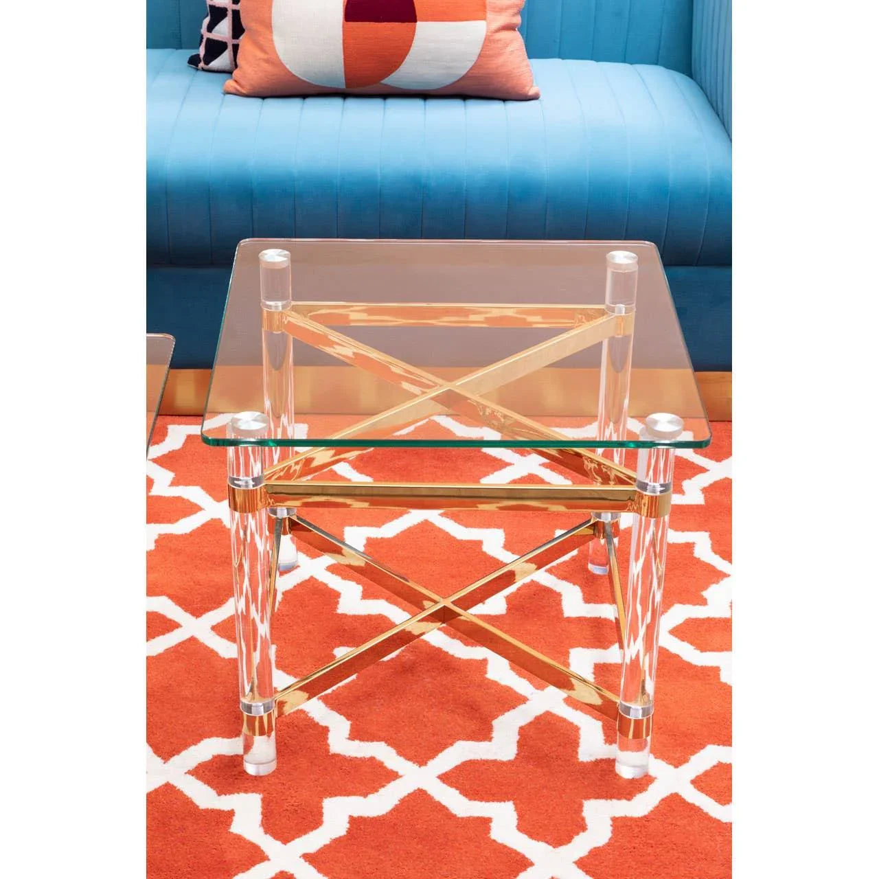Glass side table with gold and acrylic legs on orange geometric rug, blue sofa behind