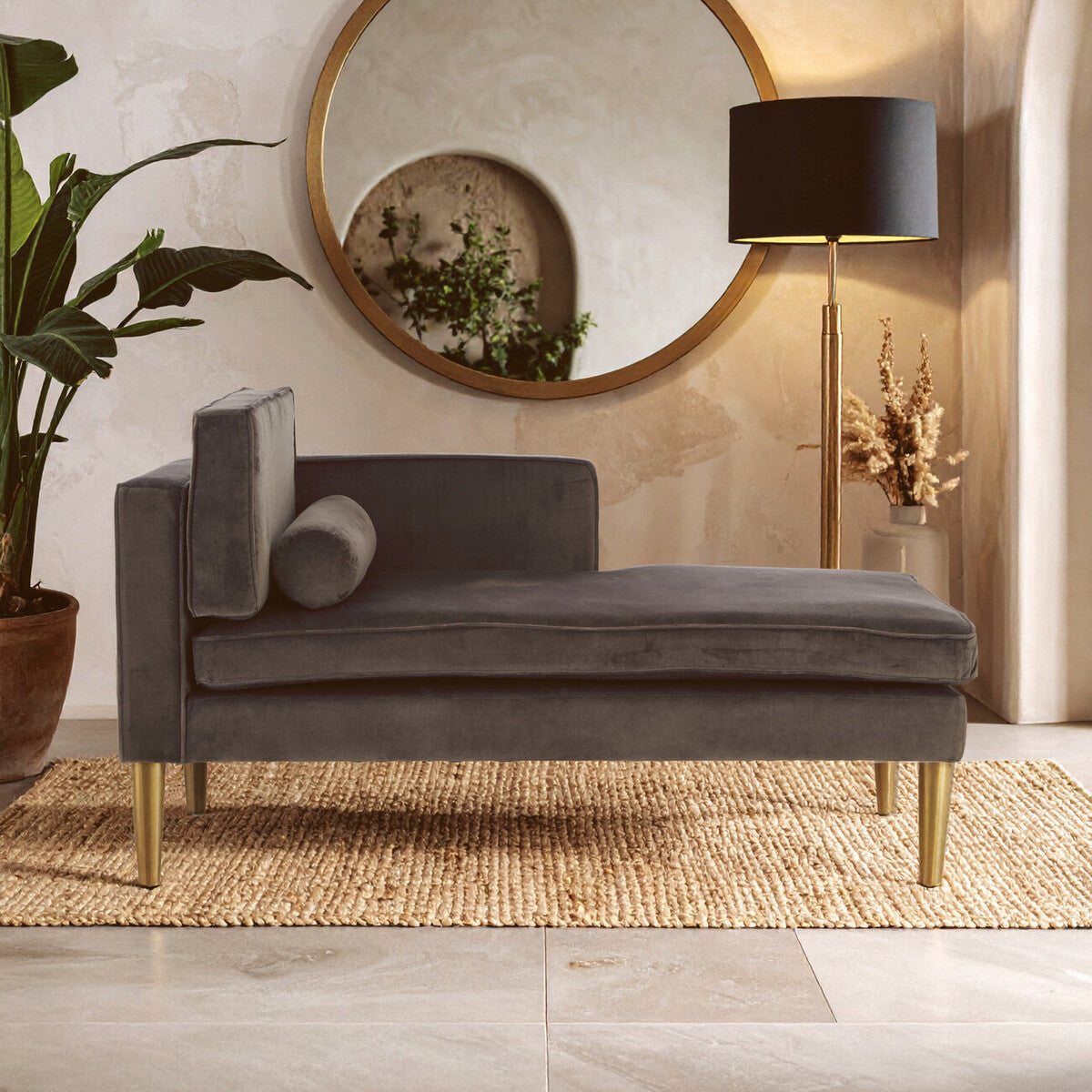 Grey velvet left-hand chaise longue with a sleek design, featuring a plush seat and a rounded backrest. A decorative cushion rests on the side, and the chaise is supported by elegant gold legs. The setting includes a large round mirror and a floor lamp, with a potted plant in the background, all placed on a textured rug