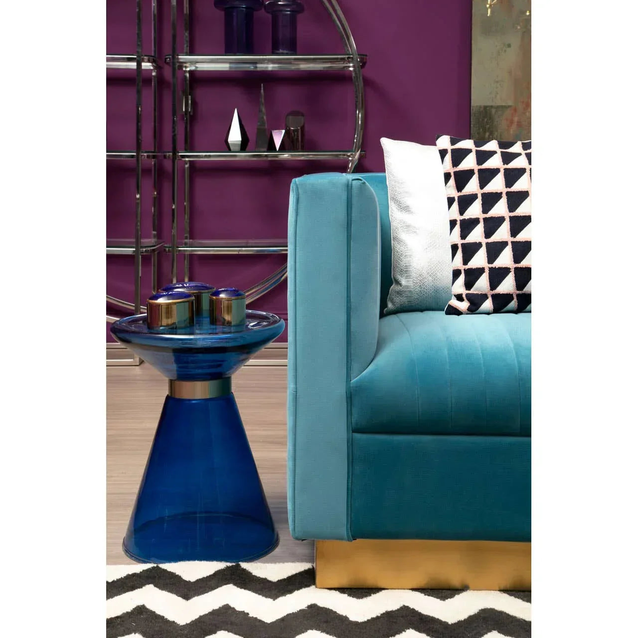 Blue glass side table beside teal velvet sofa with patterned cushions and chevron rug