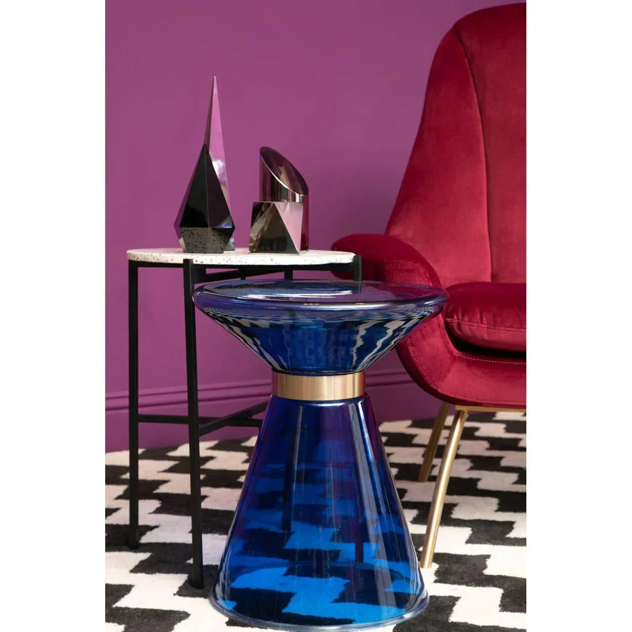 Blue glass side table beside marble accent table and red velvet armchair on patterned rug