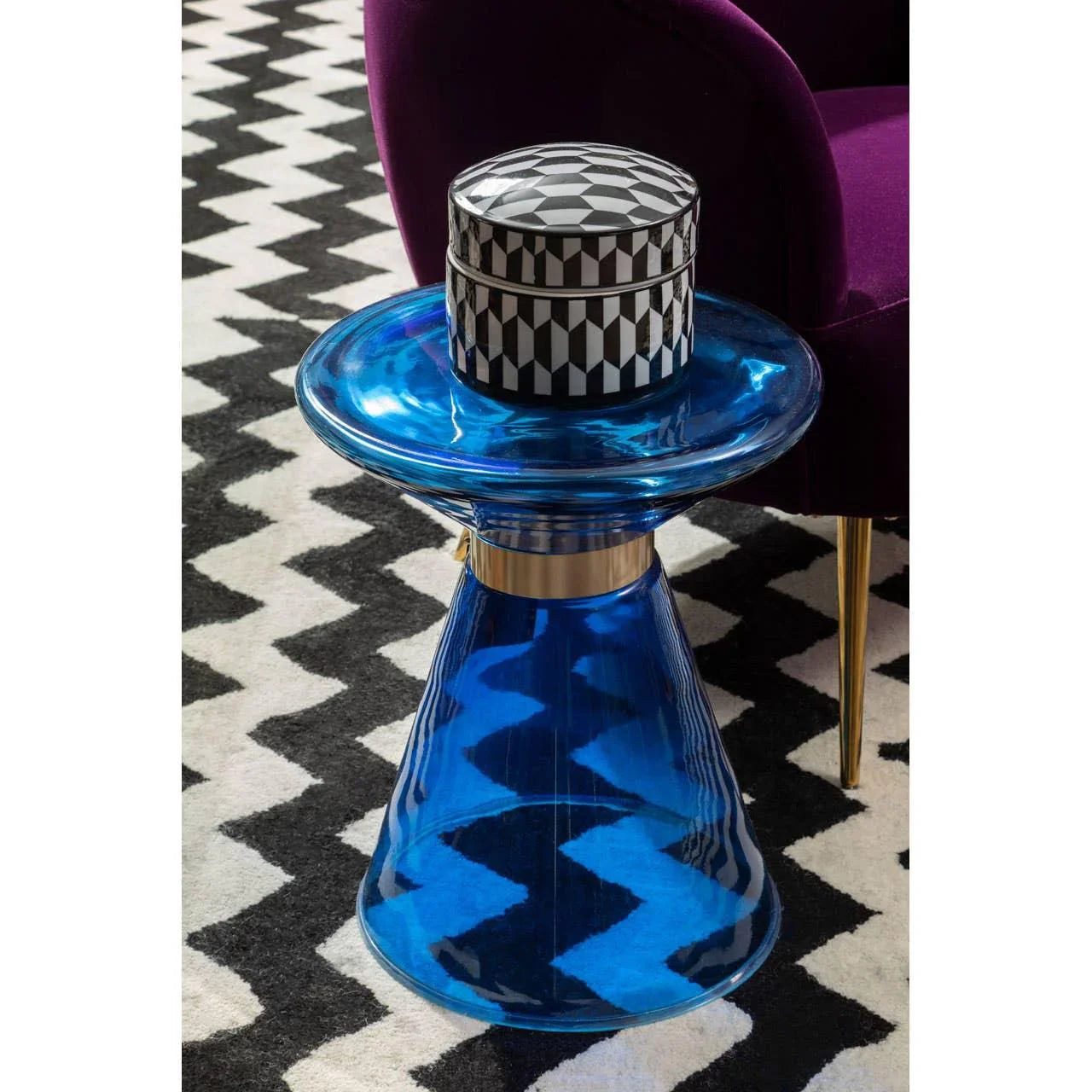 Blue glass side table with gold accent beside purple chair on black and white rug