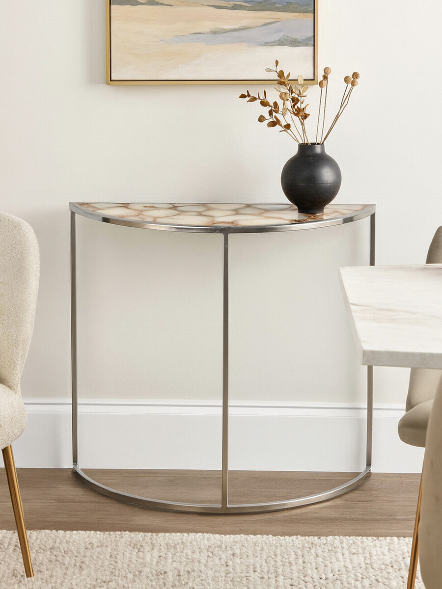 Half moon console table featuring a white agate top with natural patterns, supported by a sleek metal frame. The table is positioned against a light-colored wall, accompanied by a decorative vase and a small arrangement of dried flowers. A neutral-toned chair is visible to the left, and a portion of a dining table is seen to the right