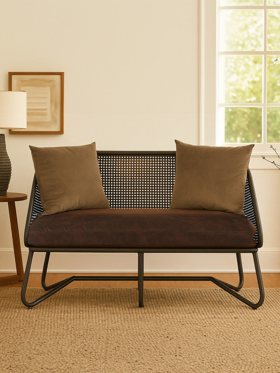 A two-seat sofa with a sleek design featuring curved legs, upholstered in a dark fabric. The sofa is adorned with two square, textured throw pillows in a complementary color. It is positioned in a cozy room with natural light coming through a window, and a light-colored rug is visible beneath it. A lamp and framed artwork are in the background, adding to the inviting atmosphere