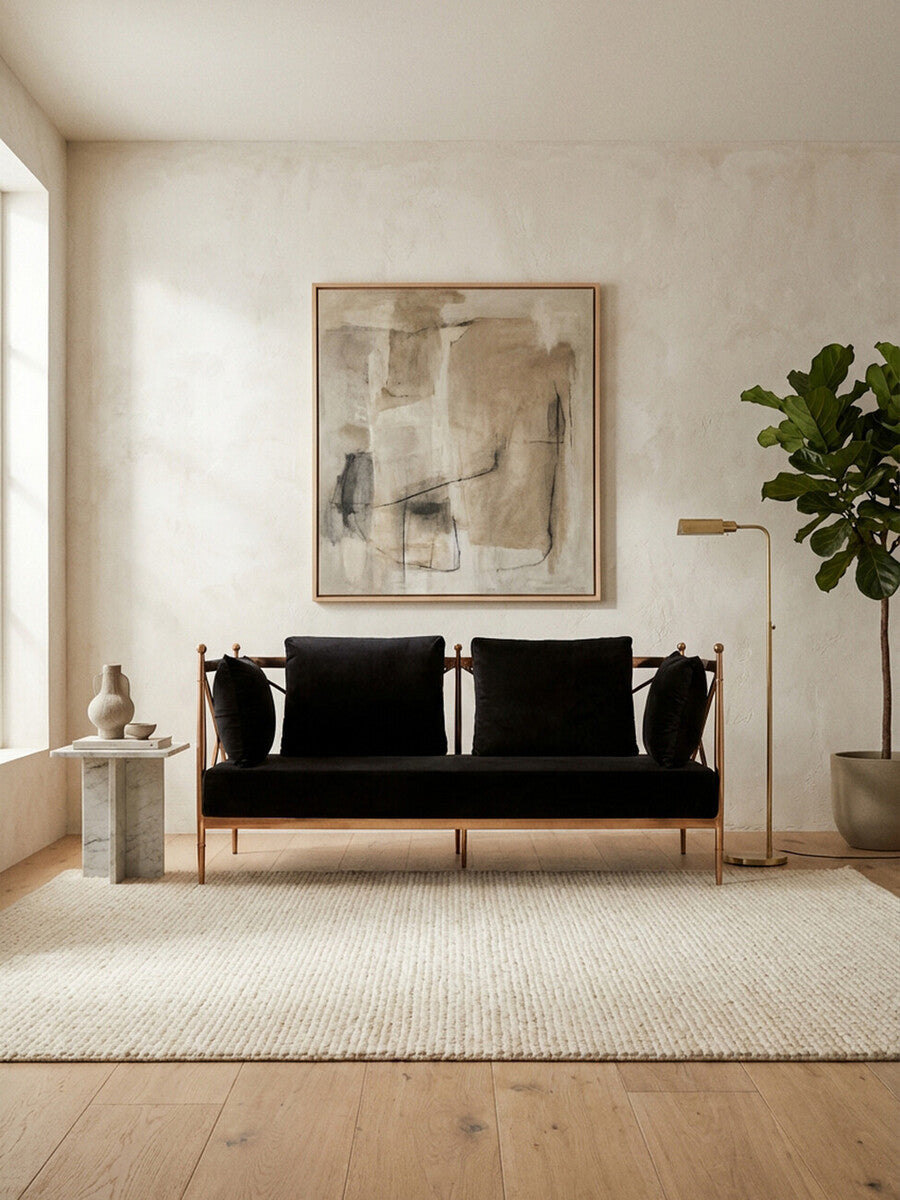 A stylish two-seater sofa upholstered in black velvet, featuring a sleek rose gold frame. The sofa is positioned in a bright room with a large window, a textured area rug beneath it, and a decorative plant nearby. A modern artwork hangs on the wall behind the sofa, adding a contemporary touch to the space