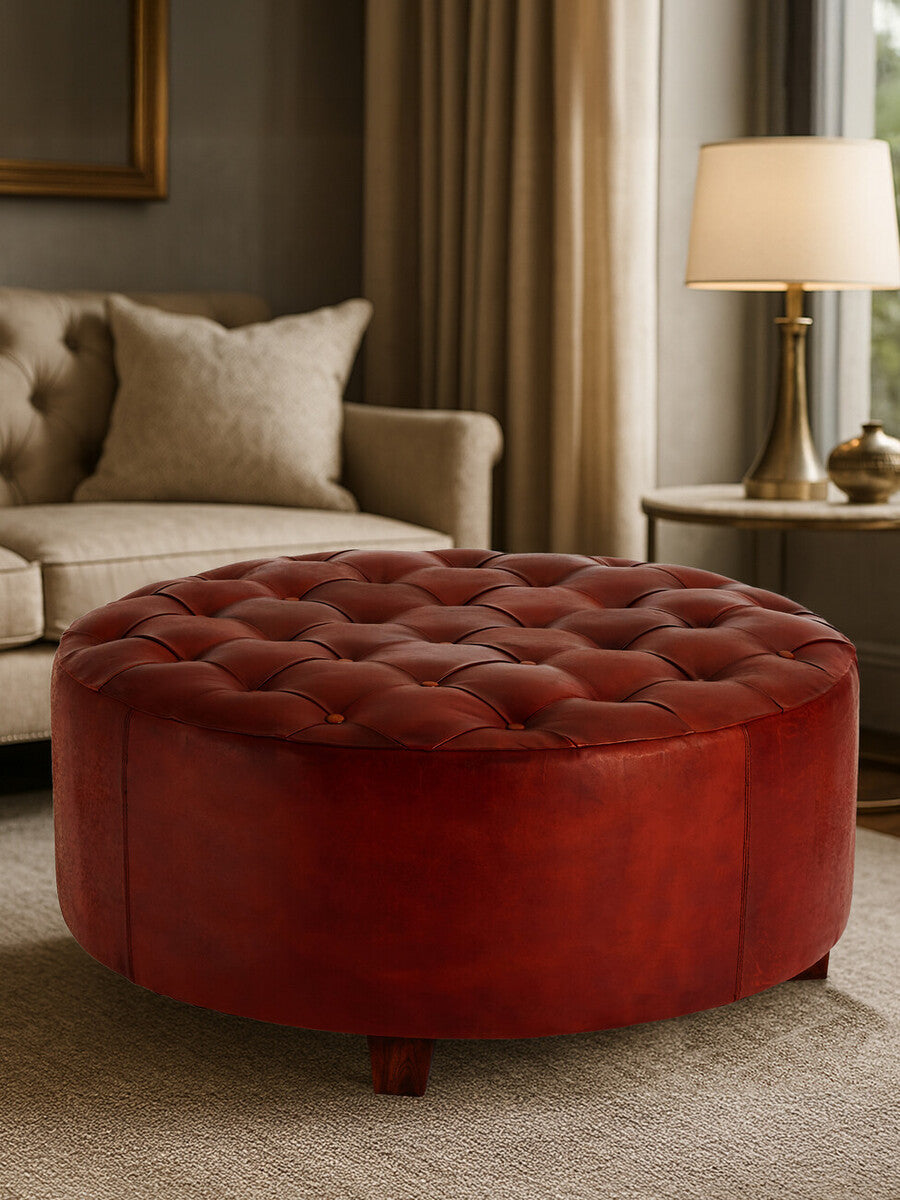 Round button tufted stool in antique brown leather, featuring a plush top with deep button detailing and a smooth, rounded base. The stool is positioned in a cozy living room setting, with a soft rug beneath it and a warm lamp in the background, creating an inviting atmosphere
