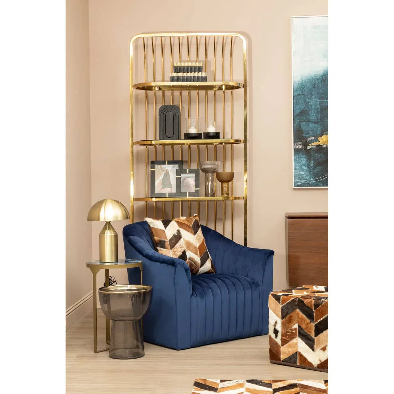 Blue velvet armchair with chevron cushion beside gold shelving unit and side table lamp