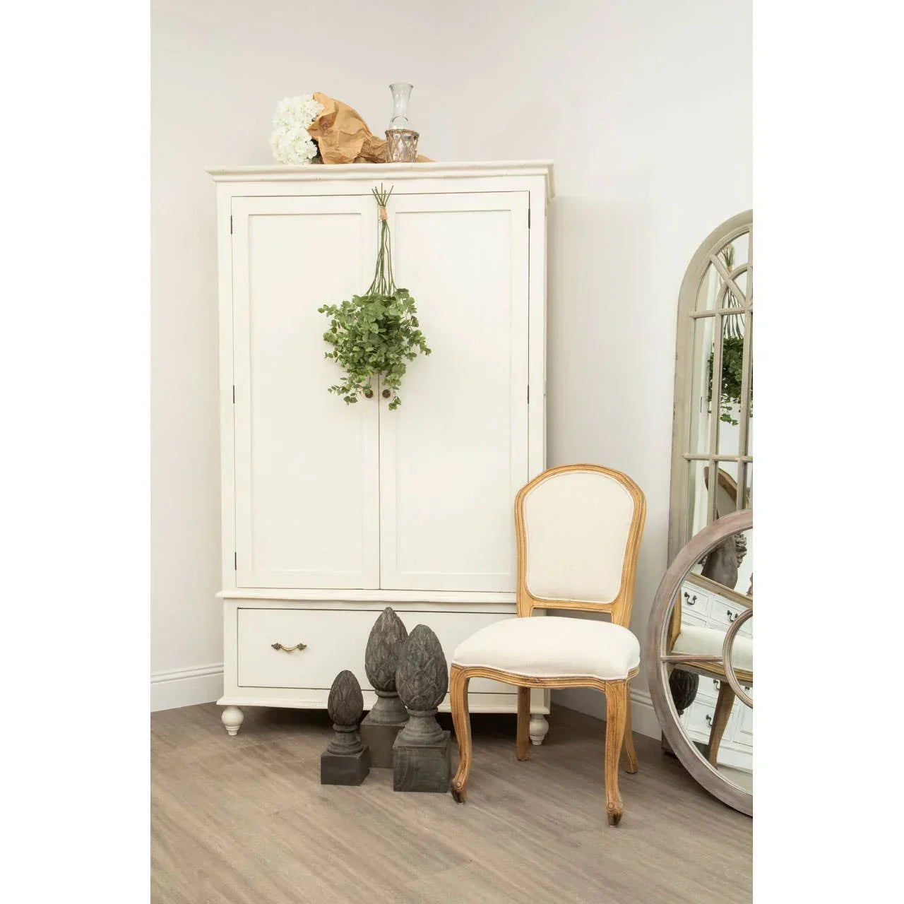 Cream wooden wardrobe with drawer beside cream upholstered dining chair and decorative mirror