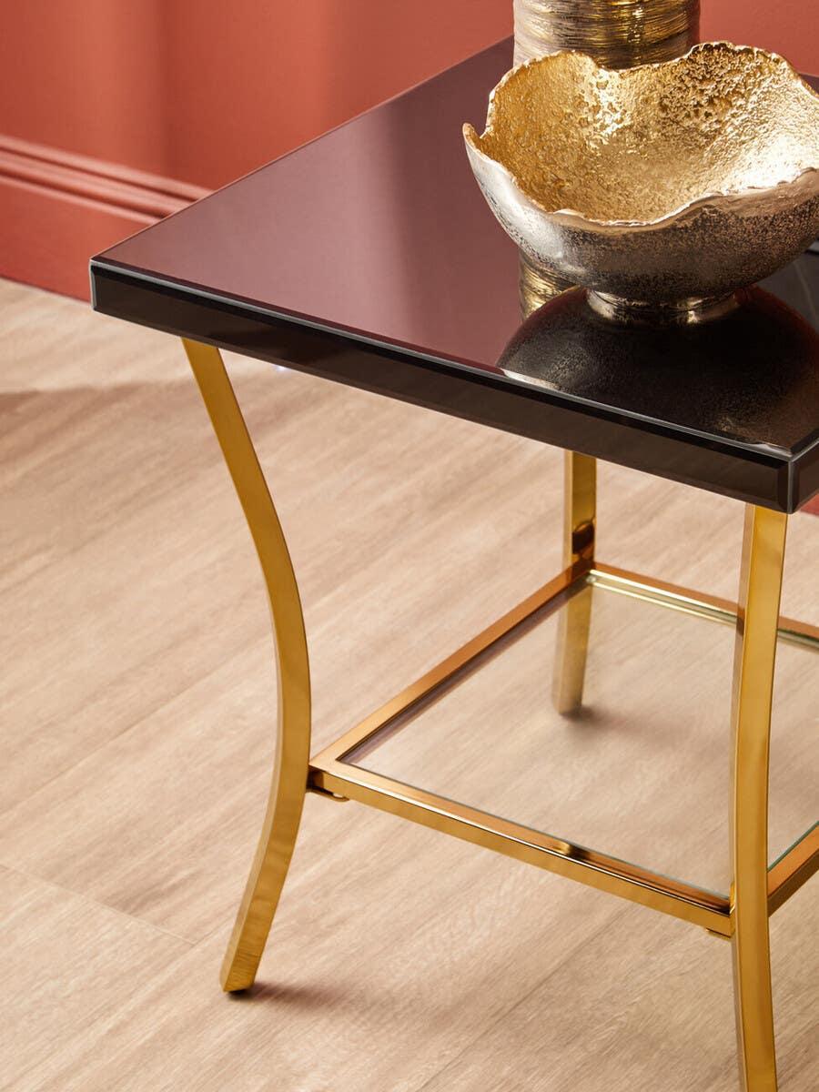Gold metal side table with black glass top and decorative gold bowl on wooden floor