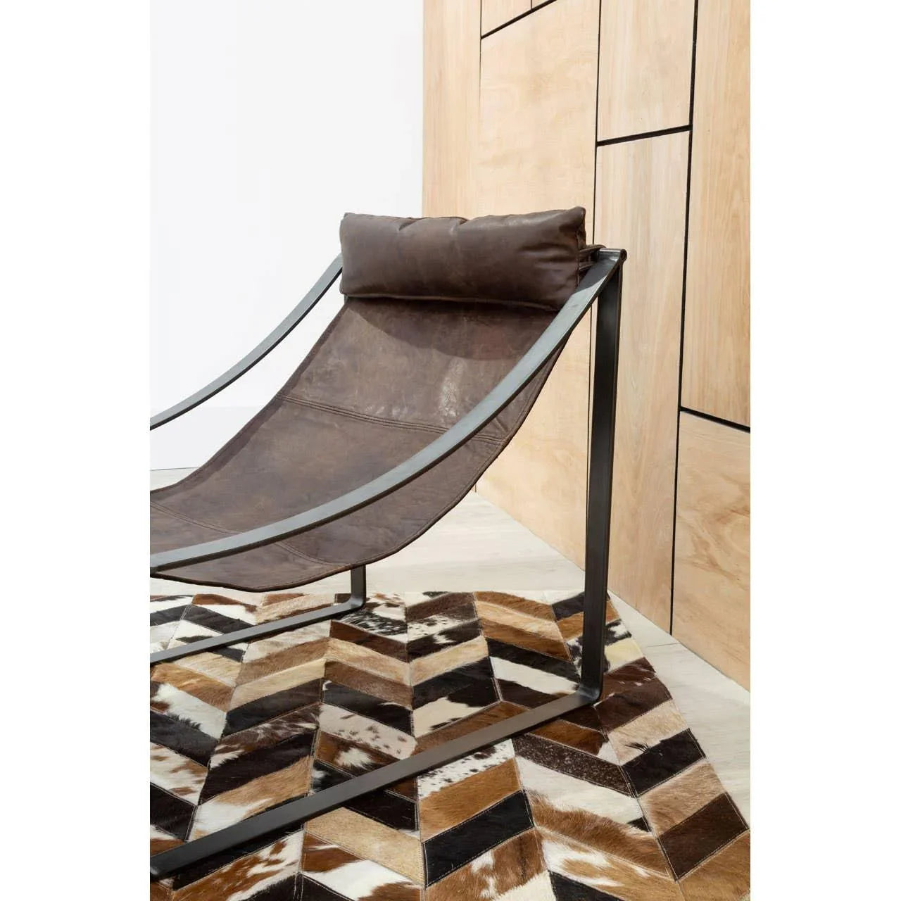 Brown leather lounge chair with metal frame on geometric cowhide rug by wood panel wall