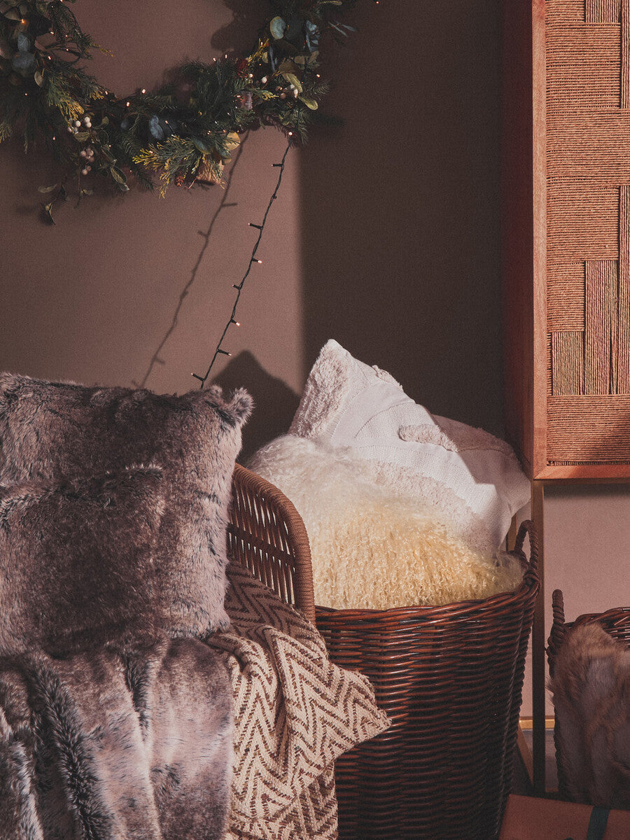 A stylish three-seater sofa featuring woven rope detailing, adorned with plush throw pillows in various textures. The sofa is positioned against a warm-toned wall, complemented by a wooden cabinet and a decorative wreath in the background, creating a cozy and inviting atmosphere
