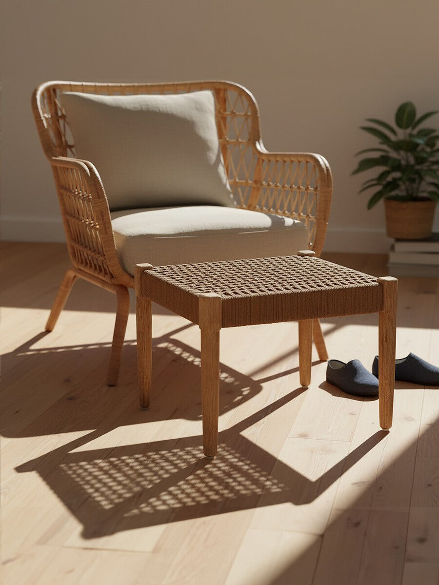 Woven rope footstool with a square shape, featuring a natural wood frame and a textured surface, placed next to a light-colored armchair with a cushion. The setting includes a warm wooden floor and a potted plant in the background, illuminated by soft natural light