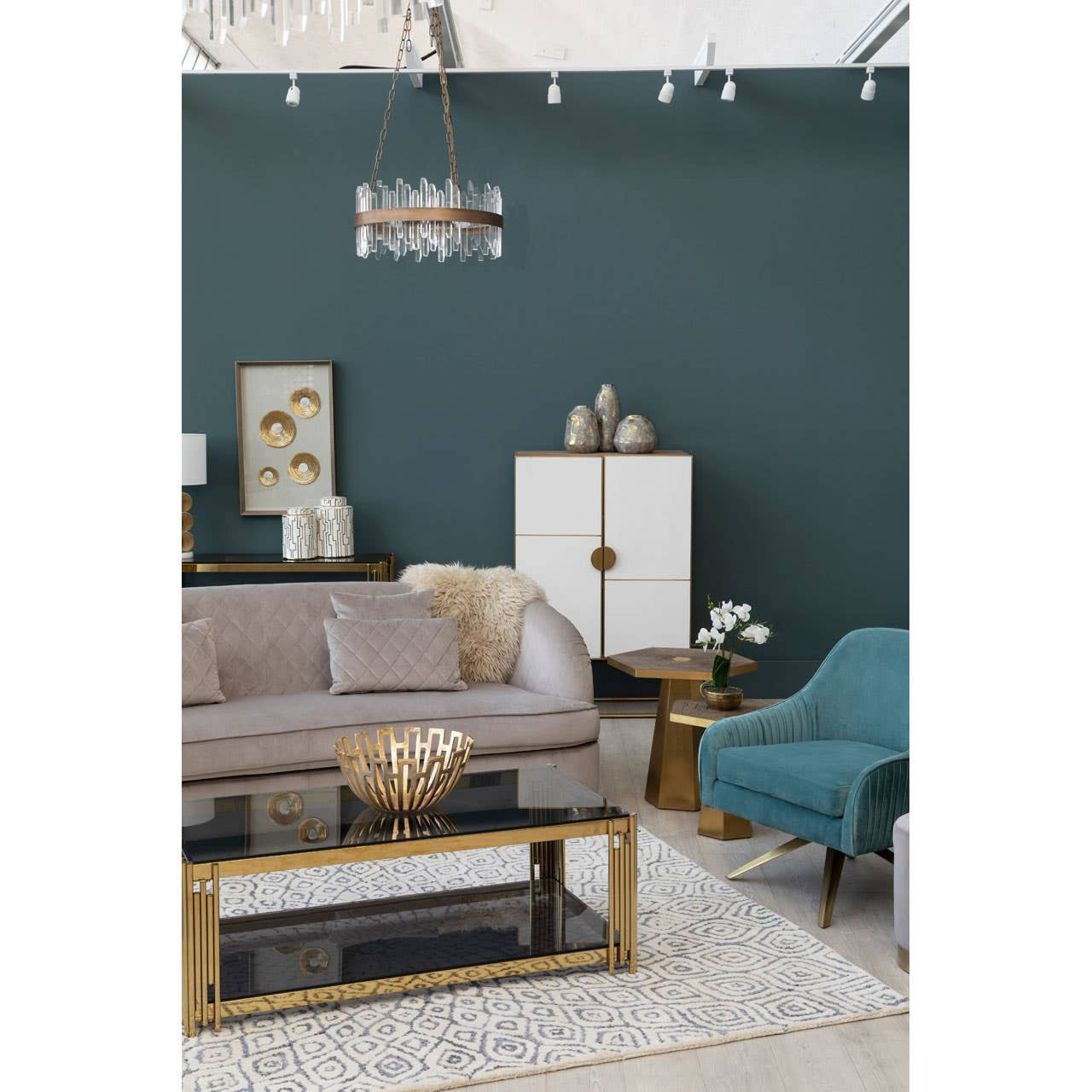 A stylish living room featuring a Modica Oak Wood and Brushed Gold Cabinet, positioned against a dark green wall. The cabinet has a sleek design with gold accents and is flanked by a light gray sofa and a teal accent chair. A round coffee table with a gold base sits in front of the sofa, and decorative items are placed on the cabinet and surrounding surfaces