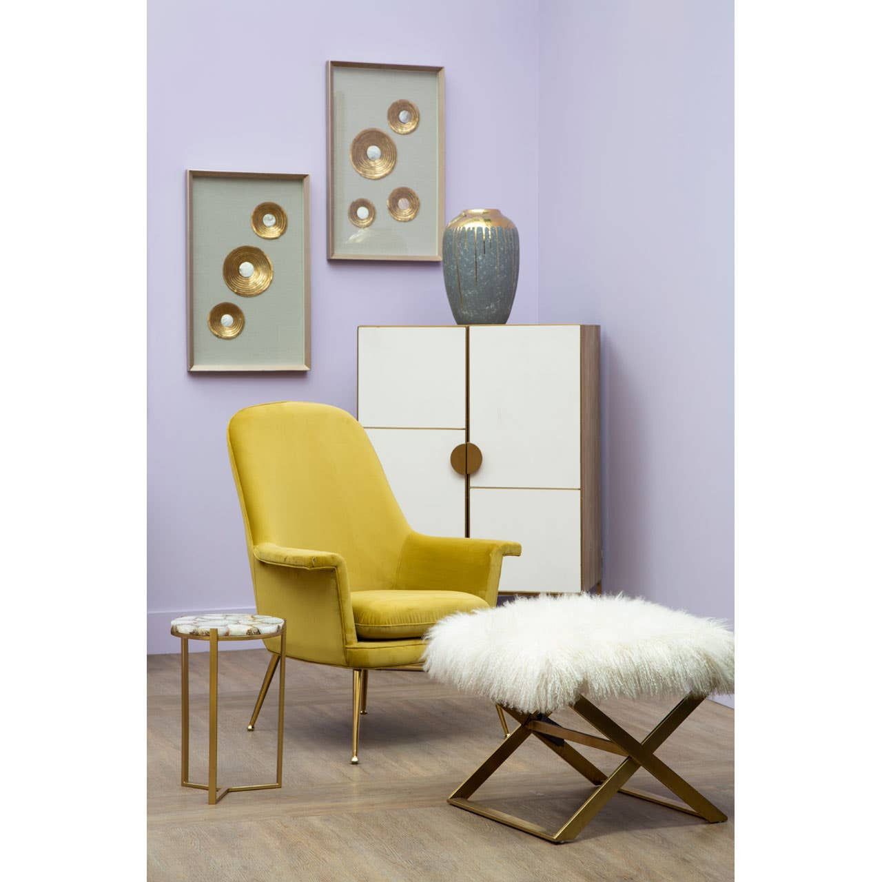 A stylish living space featuring a Modica Oak Wood and Brushed Gold Cabinet against a light purple wall. The cabinet has a sleek design with a central gold handle and two doors. A yellow armchair with gold legs sits beside a small white fur ottoman. Above the cabinet, two framed artworks with circular designs are displayed, and a gray vase is placed on top of the cabinet