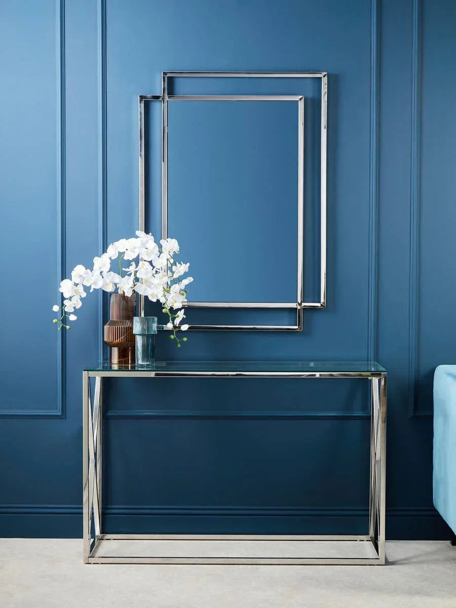 Glass console table with chrome frame, modern wall mirror, and vase against blue wall
