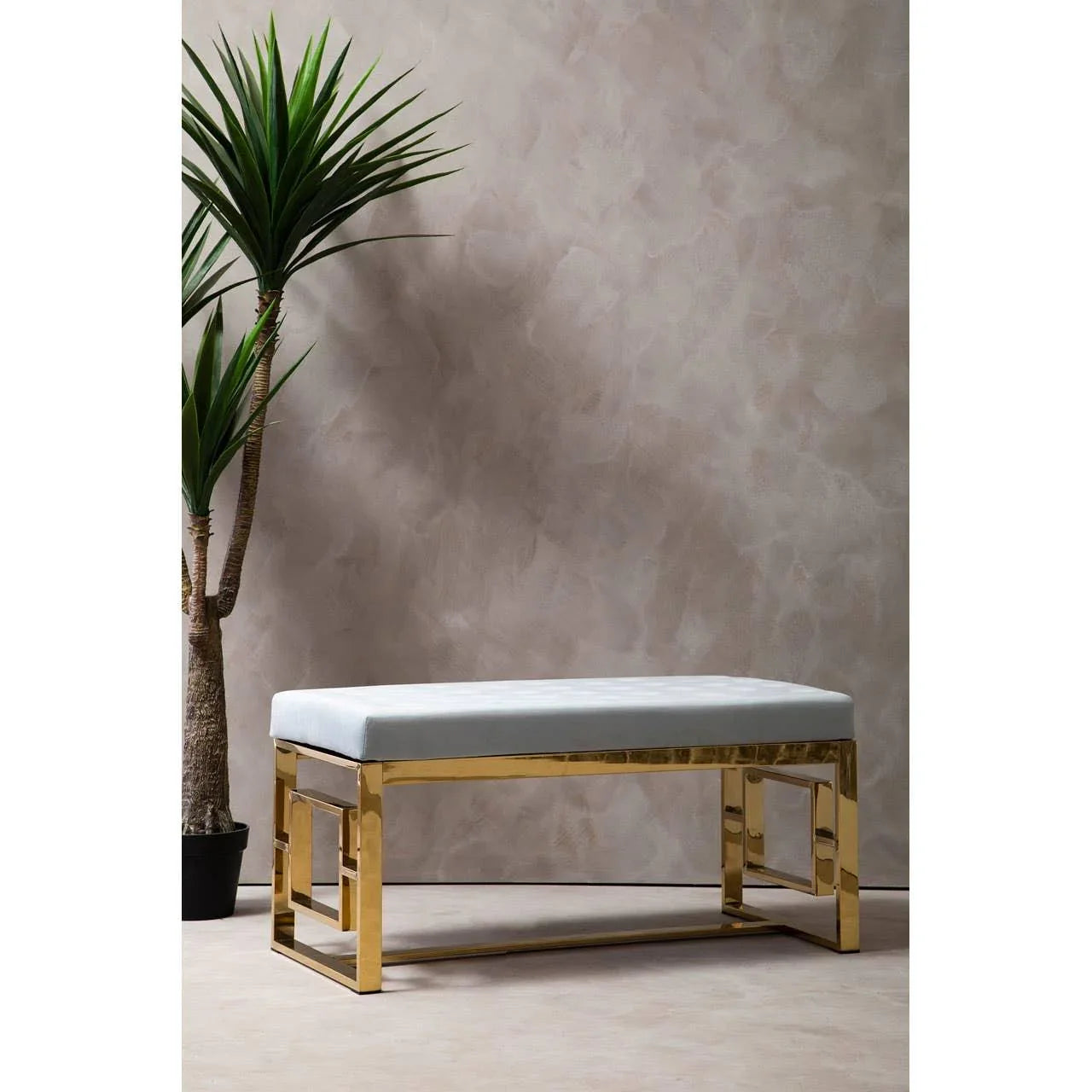Gold metal bench with white cushion beside indoor plant against textured wall