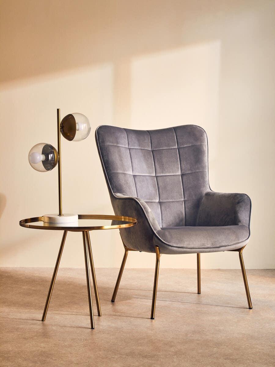 Grey velvet accent chair with gold legs next to round gold side table and modern lamp
