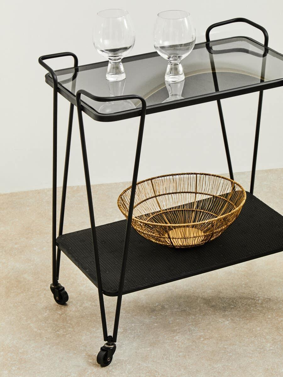 Black metal and glass bar cart with two glasses and gold wire basket on lower shelf