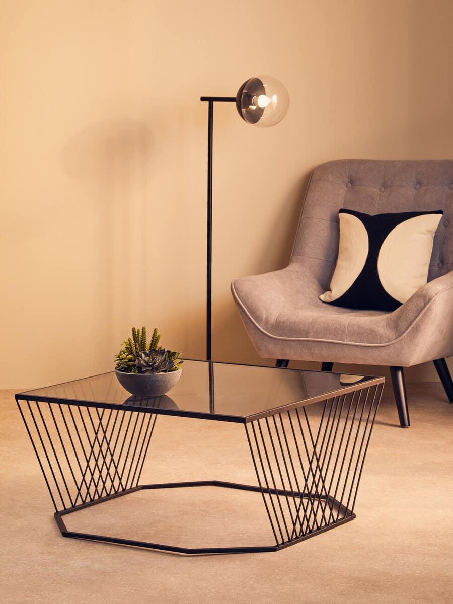 Glass top coffee table with black metal frame beside grey armchair and floor lamp