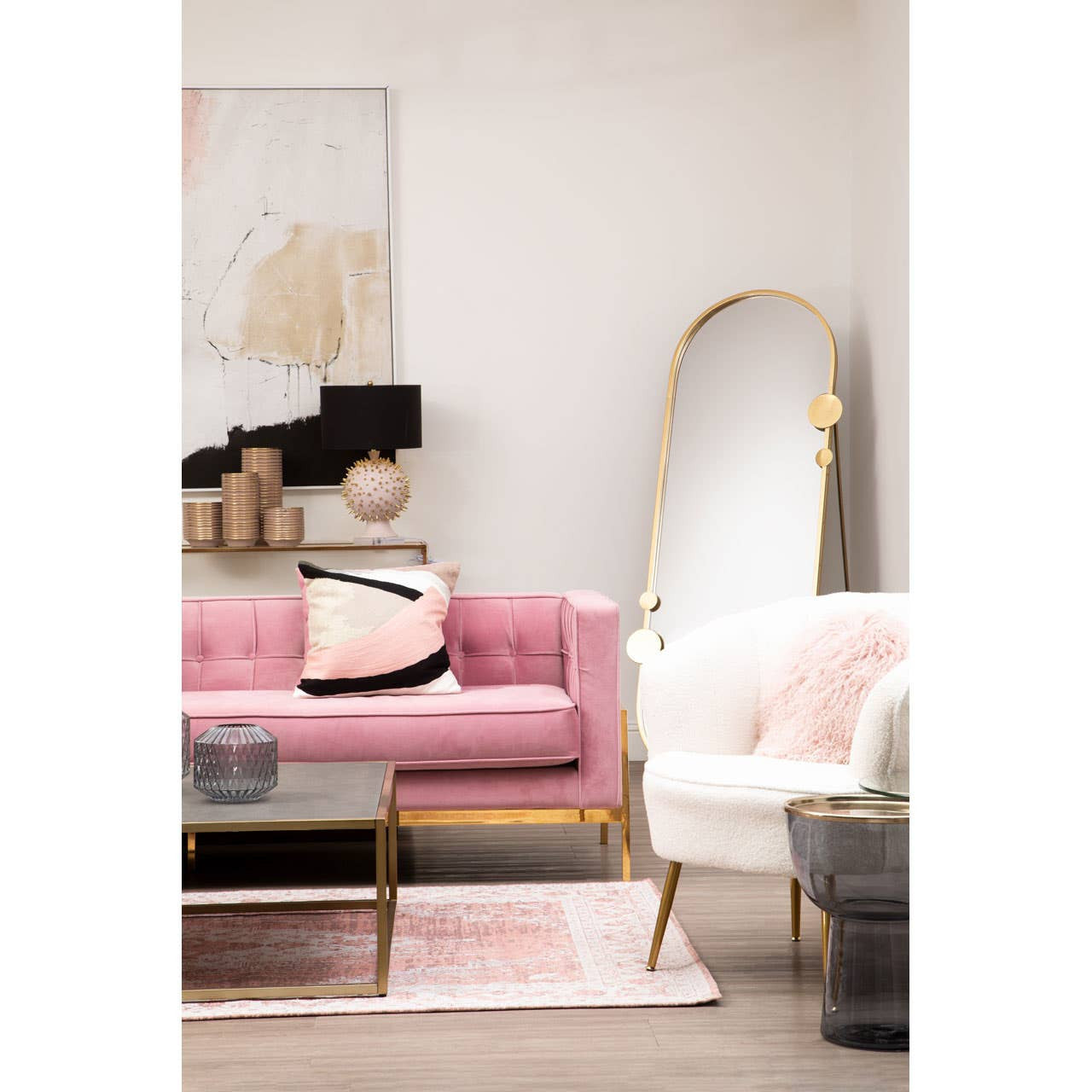 A stylish three-seat sofa in a soft pink fabric, featuring gold metal legs and a modern design. The sofa is adorned with decorative pillows and is positioned in a bright, contemporary living room setting, alongside a white accent chair and a large mirror. A light-colored rug and wall art complete the cozy atmosphere