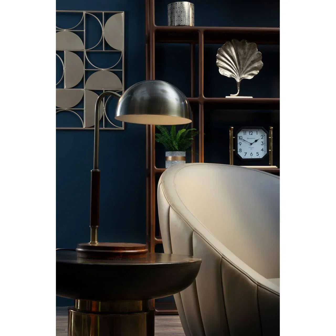 Modern beige accent chair with metal table lamp beside wooden shelving and wall décor
