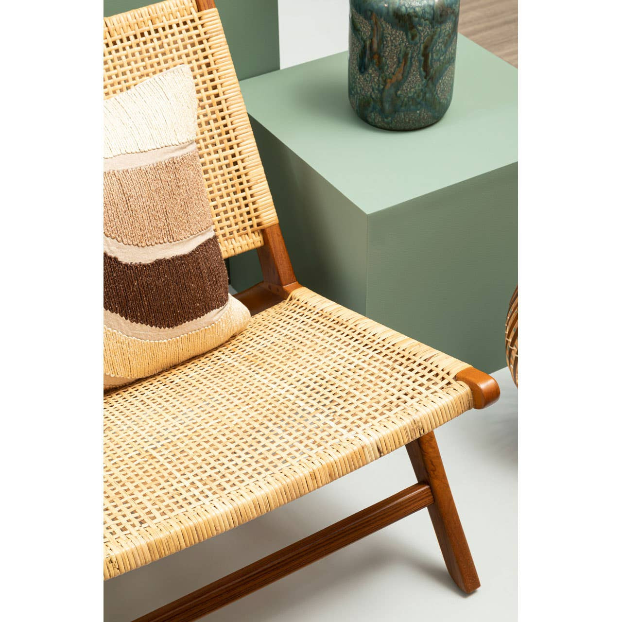 A stylish lounge chair featuring a wooden frame and natural rattan seat, complemented by a decorative cushion with a geometric pattern in neutral tones. The chair is positioned near green geometric side tables and a decorative glass vase