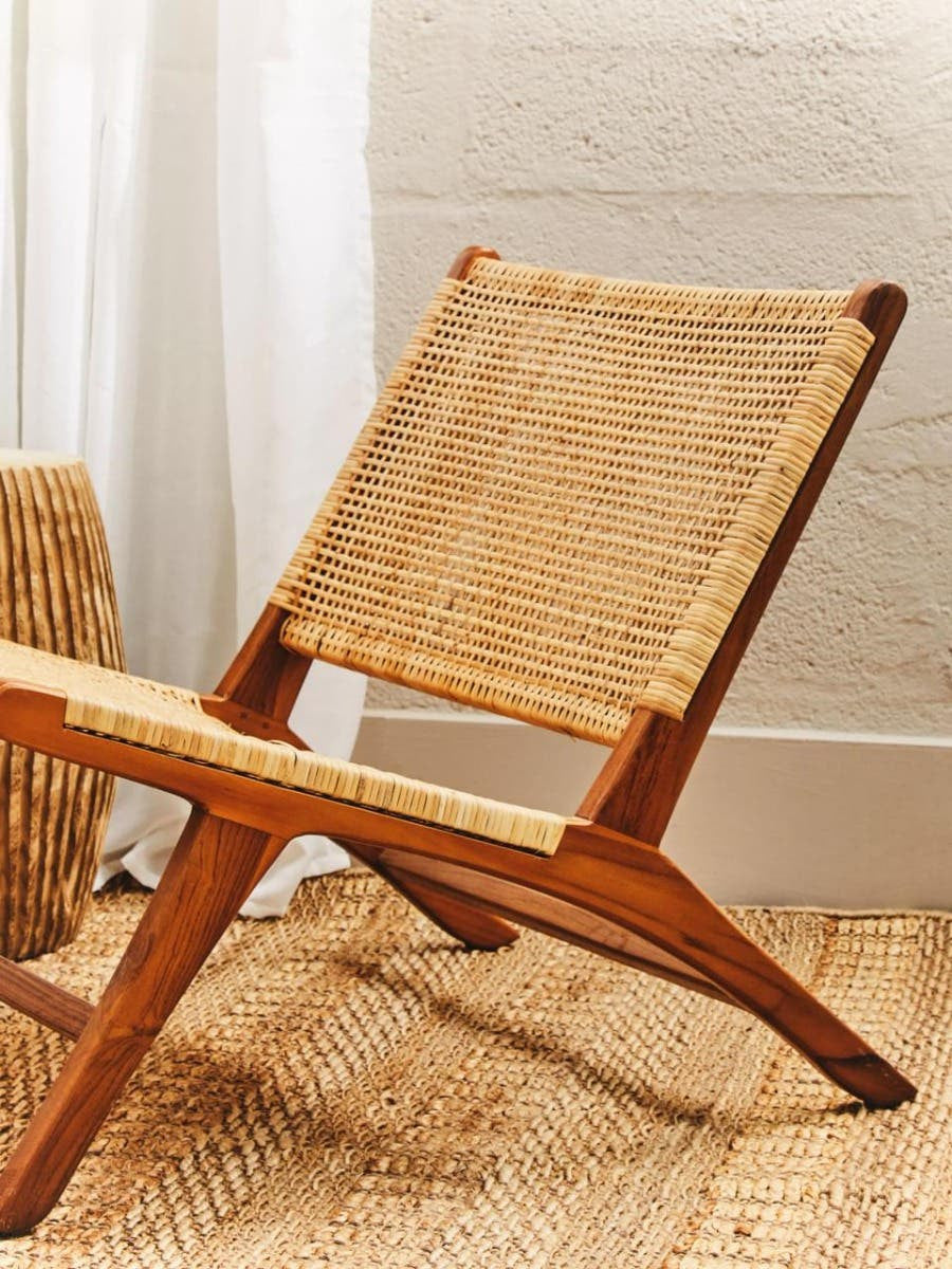 A wooden lounge chair with a natural rattan seat and backrest, featuring a minimalist design and angled legs, positioned on a textured rug with soft lighting and a light-colored wall in the background