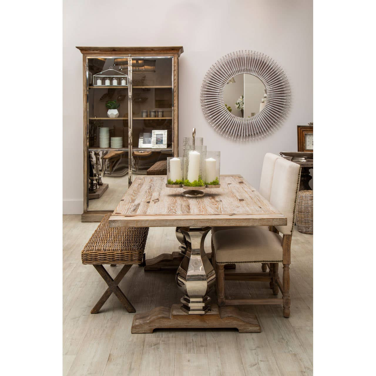 White washed pine wood cabinet with two doors, featuring a rustic design. The cabinet is placed in a dining area alongside a wooden table set with candles and decorative items. Light-colored chairs are positioned around the table, and a round mirror is visible on the wall behind the cabinet. The overall ambiance is warm and inviting, with natural wood tones and soft lighting