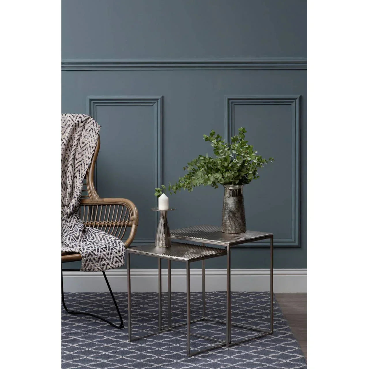 Metal nest of tables with patterned tops, candle holder, vase and plant, next to wicker chair