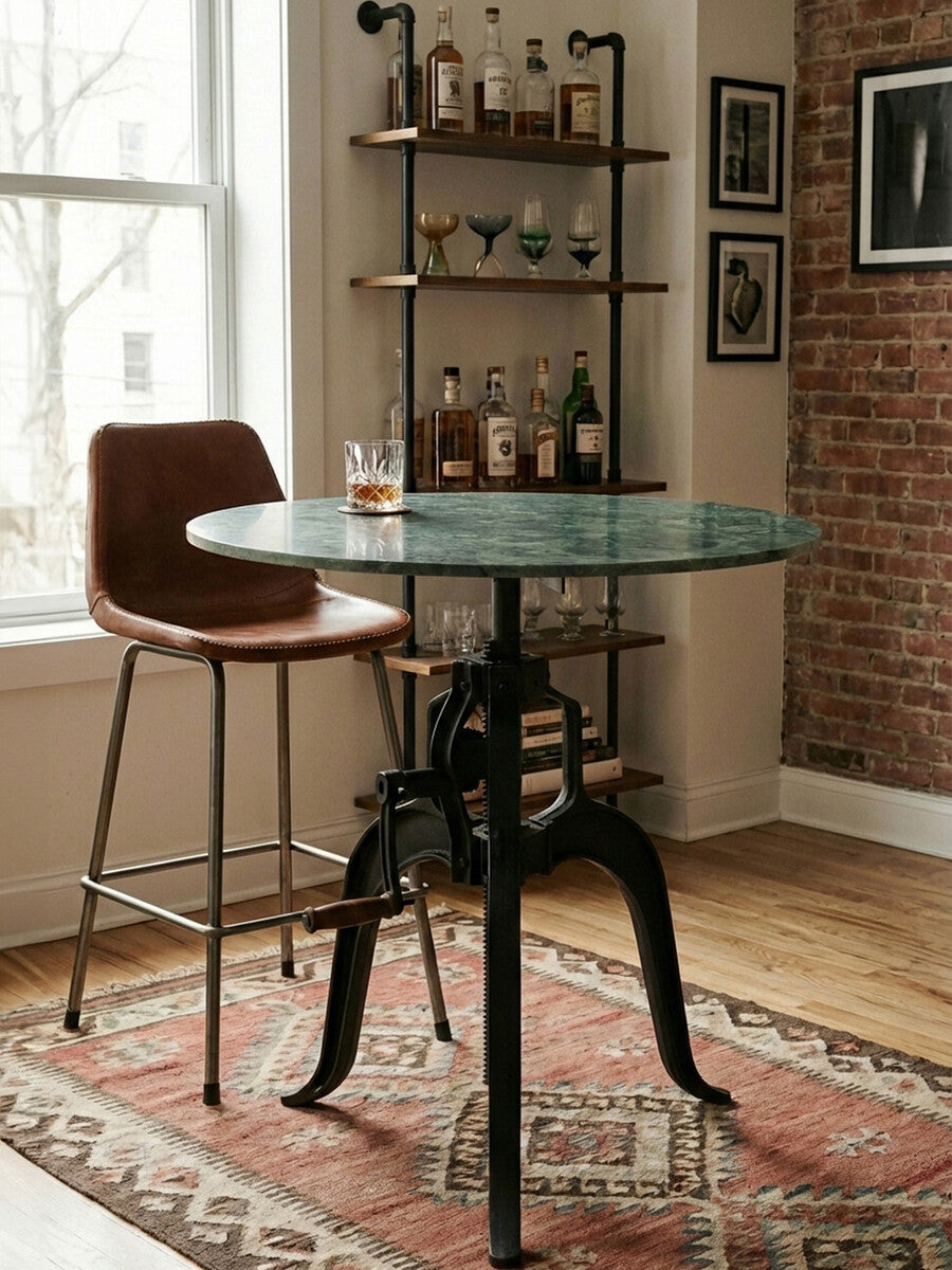 Small bar table featuring a round green marble top supported by a black metal base with intricate detailing. A brown leather bar stool is positioned next to the table, and a rustic rug is visible beneath. The background includes a brick wall and a shelf displaying various bottles and glassware. Natural light filters in through a nearby window