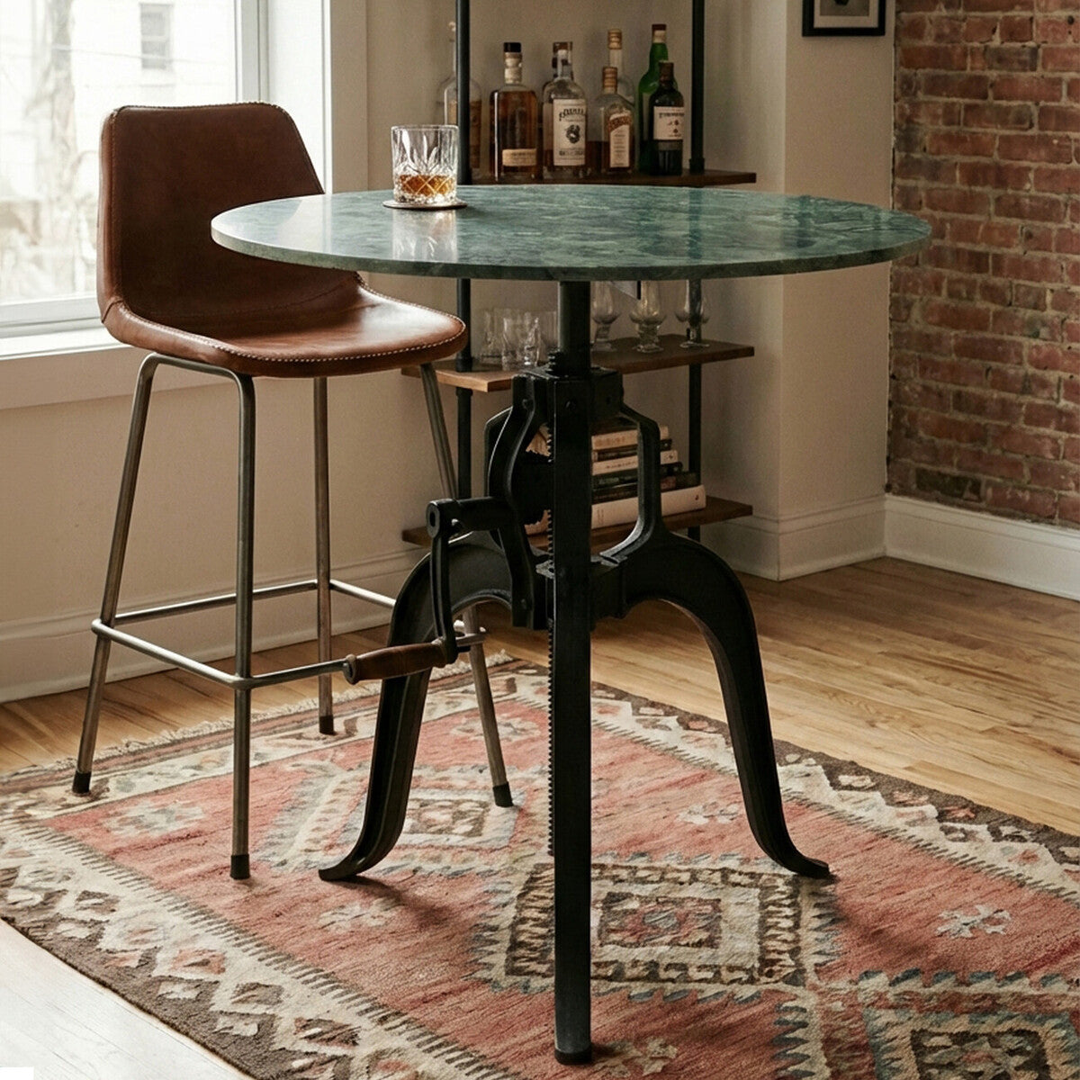 Small bar table featuring a round green marble top supported by a black metal base with a vintage design. A brown leather bar stool is positioned next to the table, and the setting includes a patterned area rug and a backdrop of exposed brick walls with a bar cart displaying bottles
