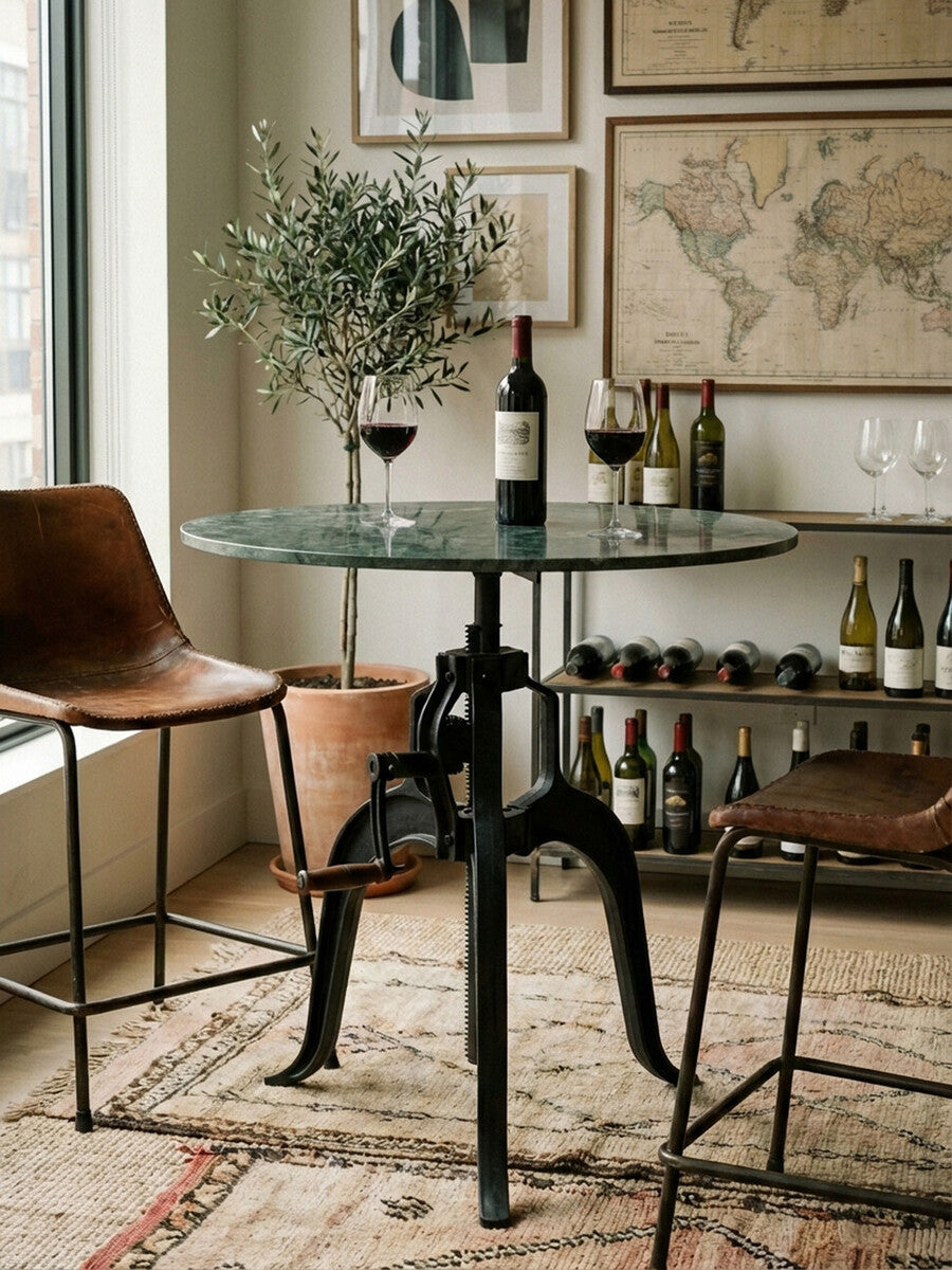 Small bar table featuring a round green marble top supported by a black metal base with a unique design. Two brown leather chairs are positioned beside the table, and a bottle of wine with two glasses sits atop the table. In the background, a collection of wine bottles is displayed on a shelf, and framed maps adorn the wall. A soft area rug is visible beneath the table and chairs, adding warmth to