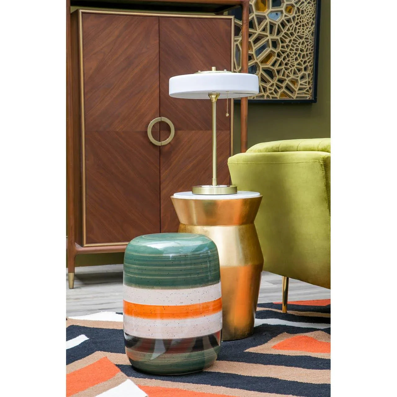 Striped ceramic side table beside gold accent table with lamp, green sofa, and wood cabinet