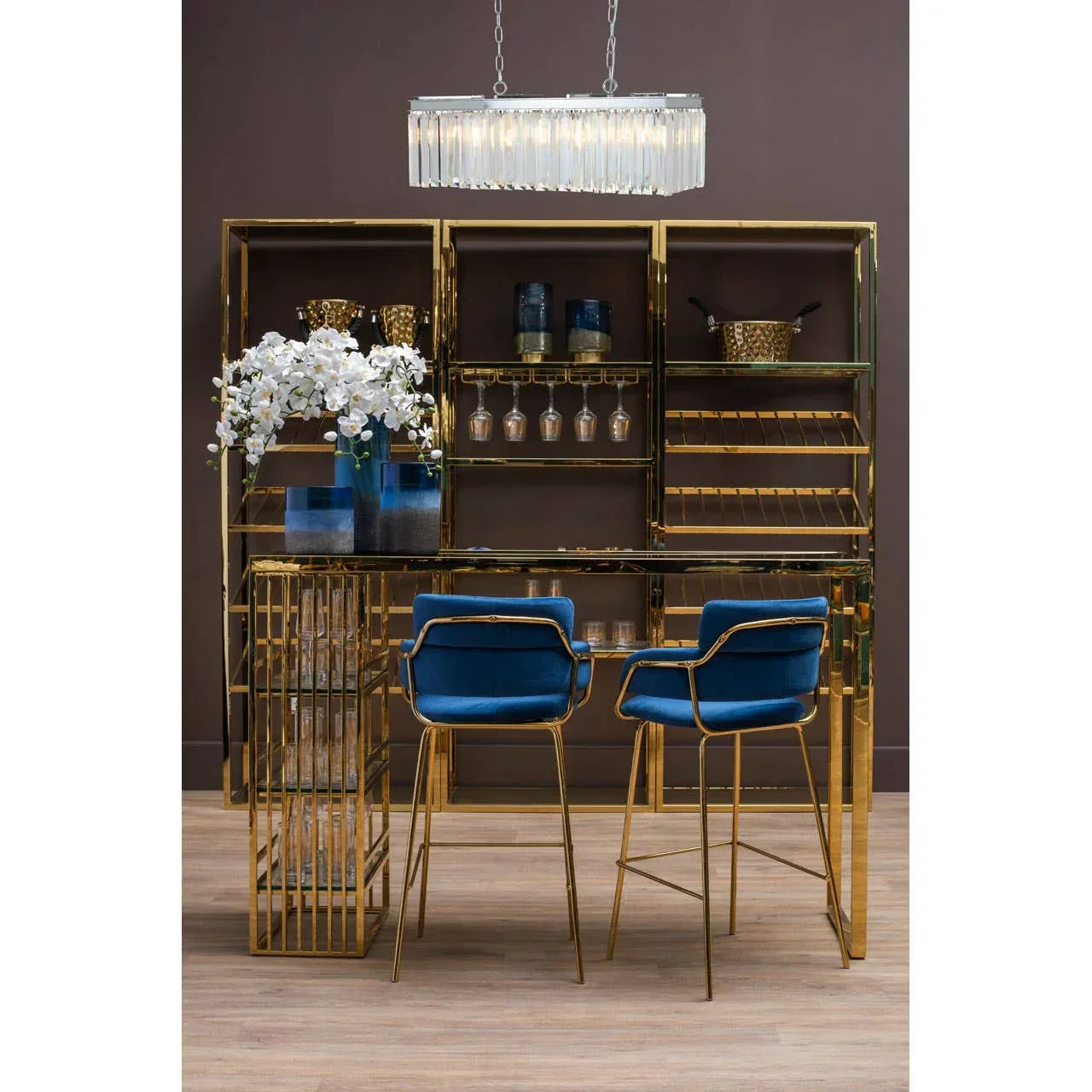 Gold home bar set with glass shelves, blue velvet bar stools, and modern pendant light