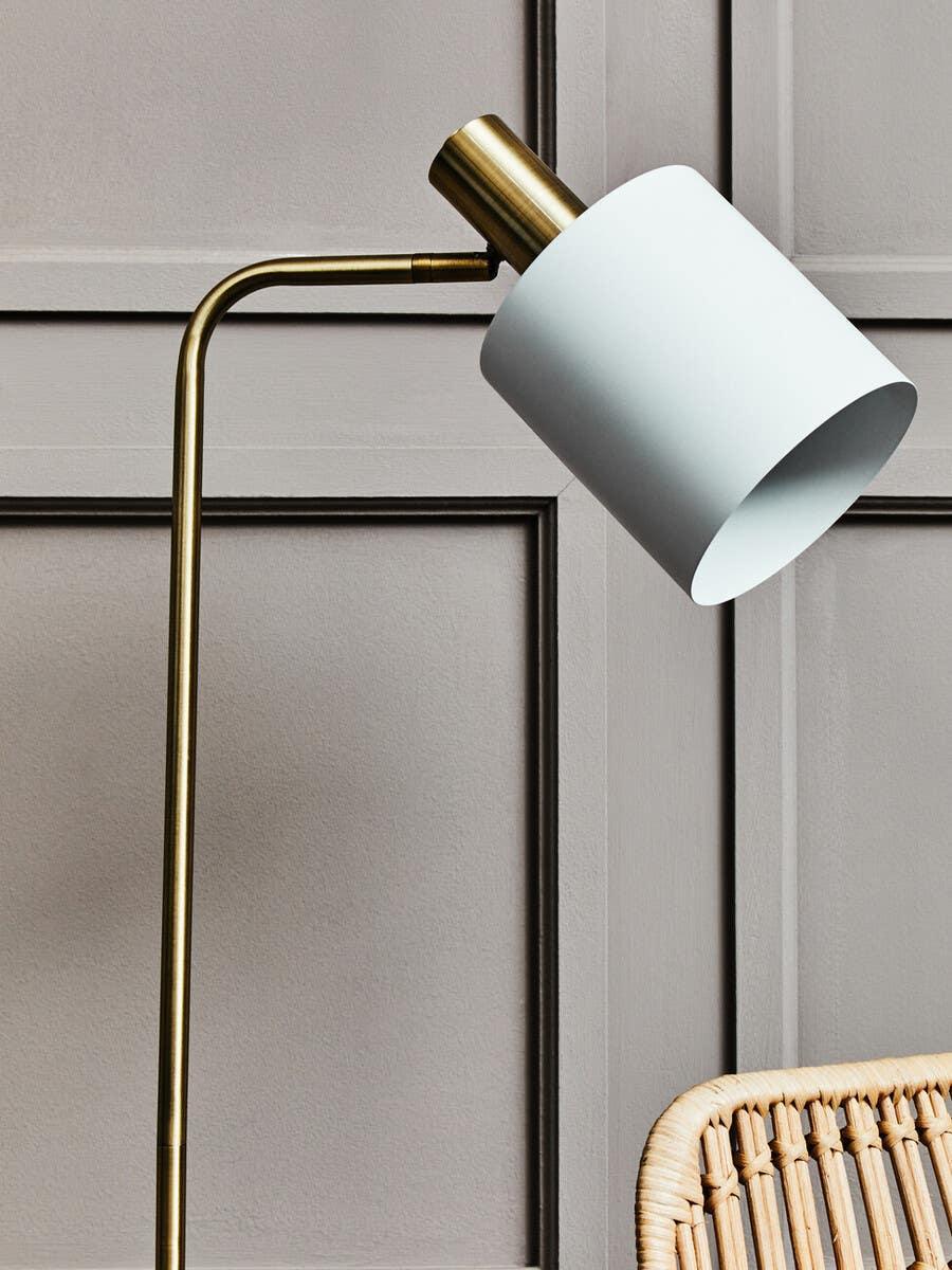 Brass floor lamp with white cylindrical shade beside rattan chair against panelled wall