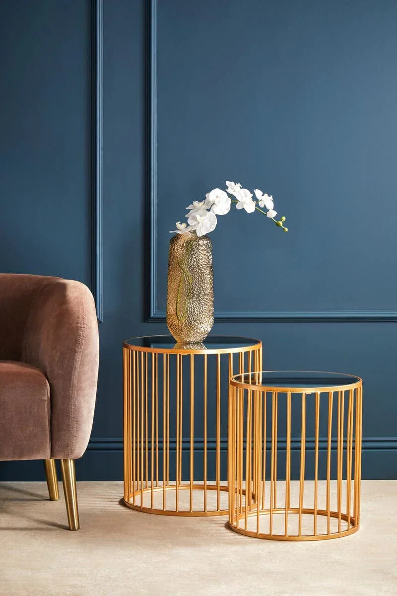 Gold metal nesting side tables with glass tops beside velvet armchair and vase with flowers