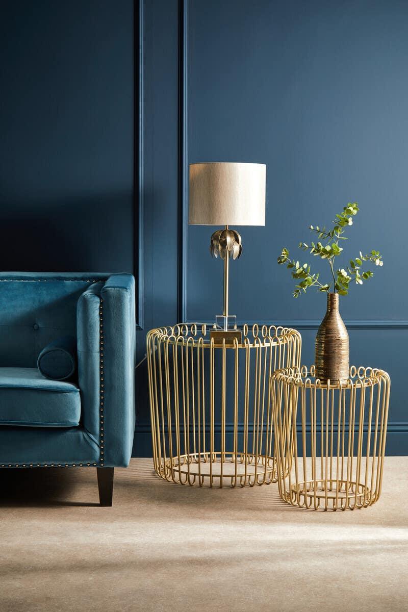 Gold wire side tables with table lamp and vase next to blue velvet sofa against blue wall