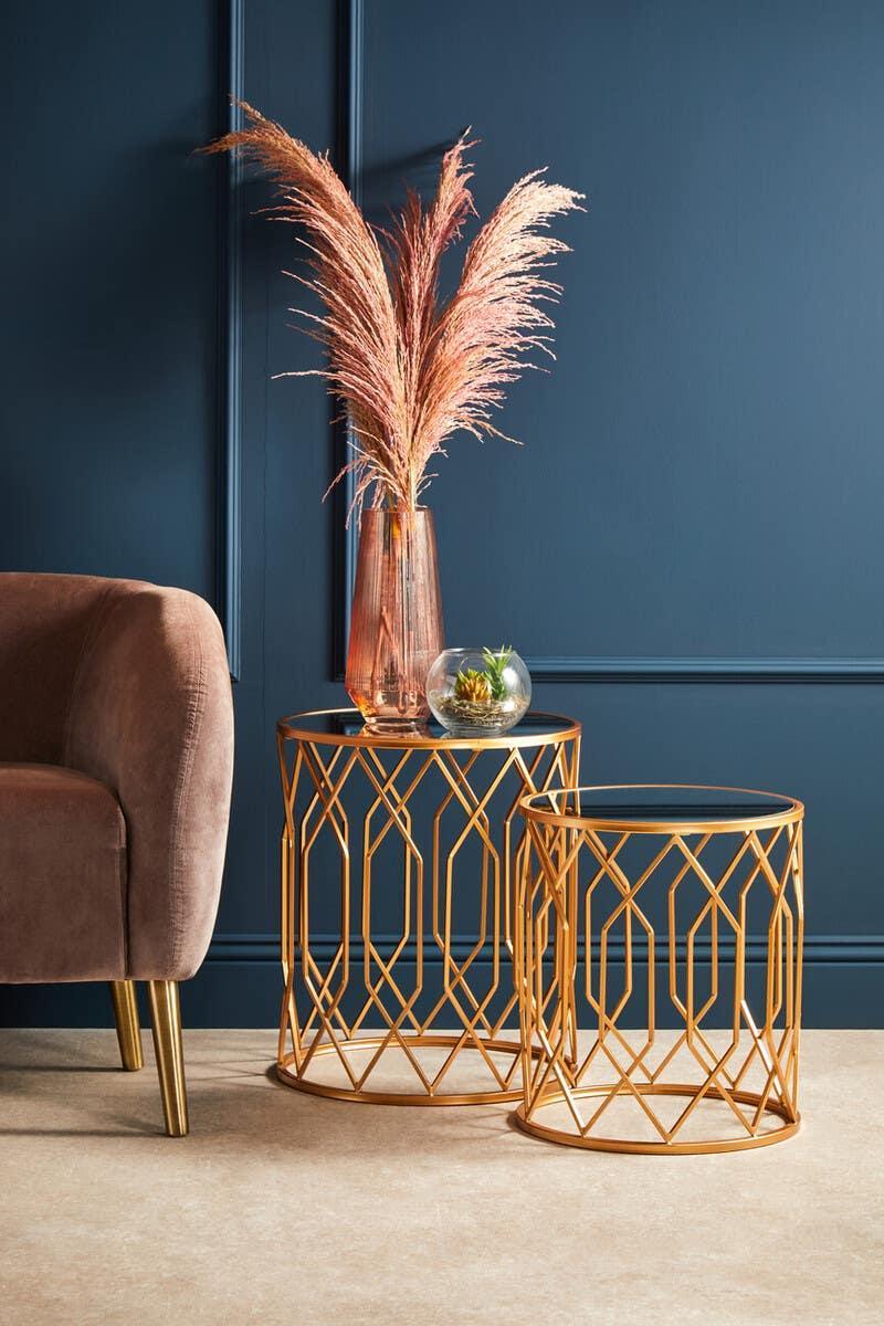 Gold metal nesting tables with glass tops beside a blush velvet armchair and decorative vase