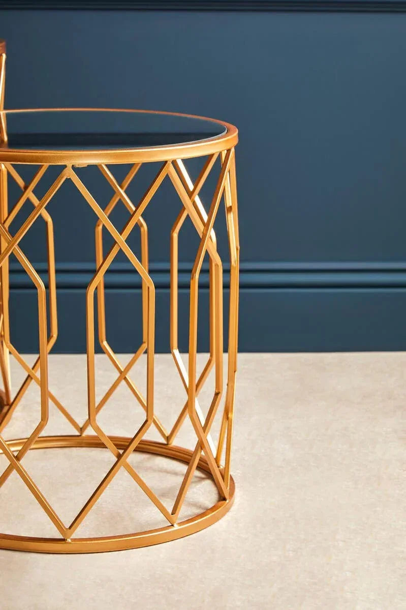 Gold metal side table with geometric frame and glass top against dark blue wall