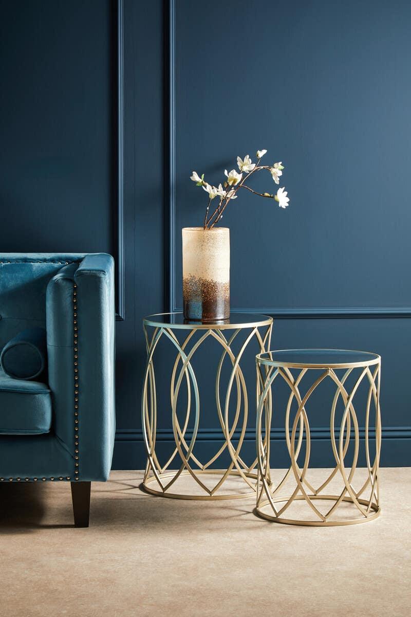 Gold metal side tables with glass tops beside teal velvet sofa and decorative vase