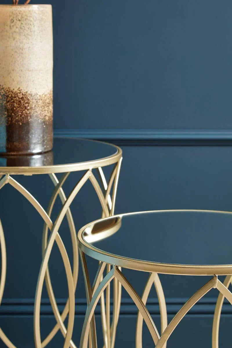 Gold metal side tables with glass tops and geometric bases against dark blue wall