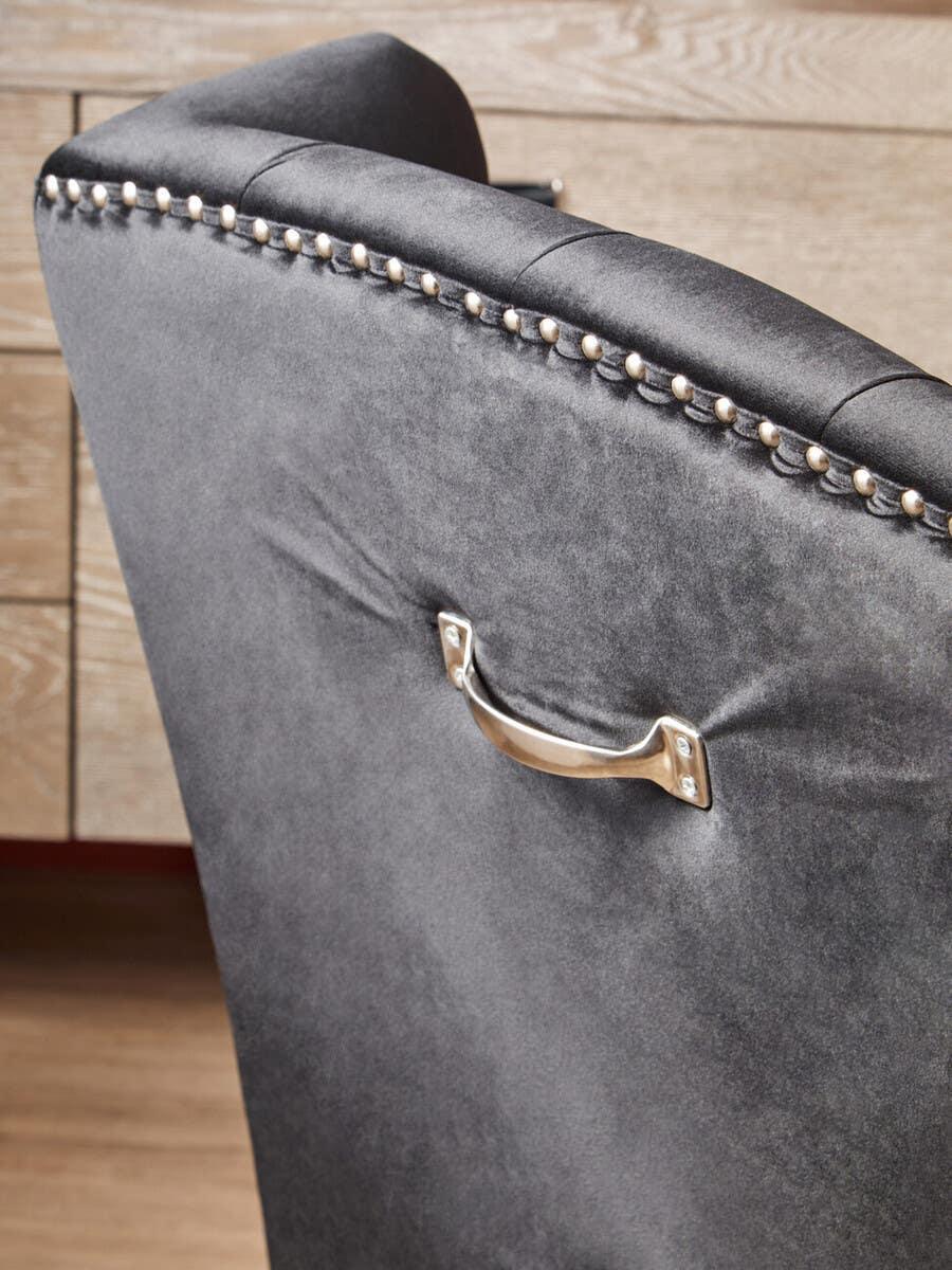 Grey velvet dining chair with chrome stud detailing and back handle against wood background