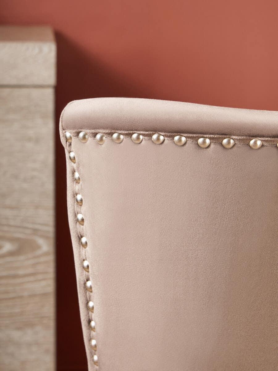 Beige velvet dining chair backrest with metal stud trim beside light wood table