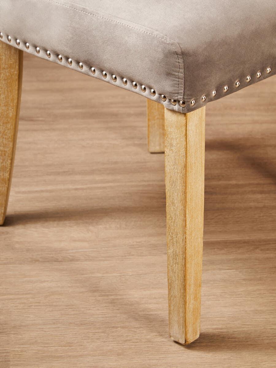 Beige fabric dining chair with wooden legs and silver nailhead trim on oak floor