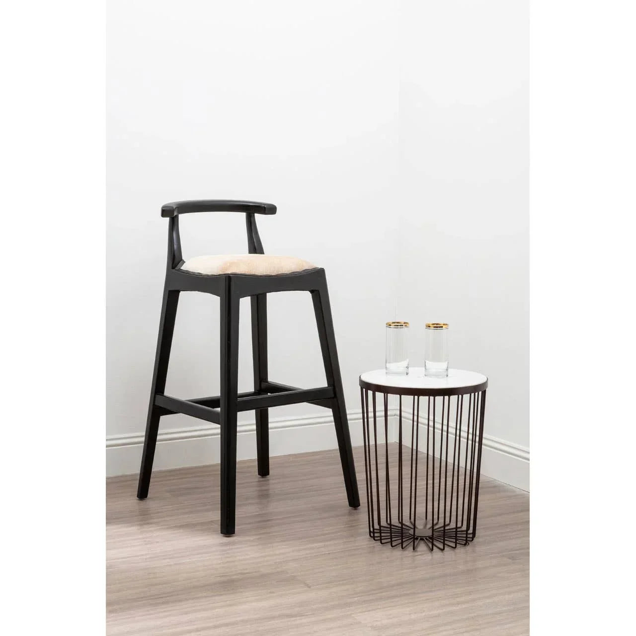 Black wooden bar stool with beige seat beside round wire side table and two glasses