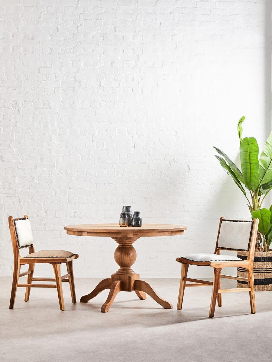 Wood dining table set with two chairs and white cushions, round base, indoor plant beside