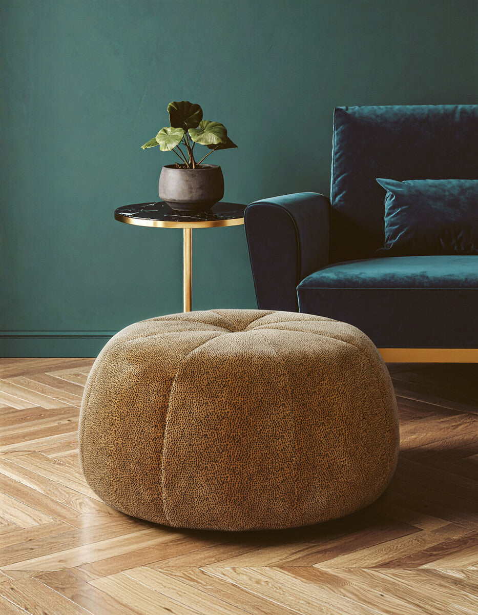 Round pouffe with a leopard print pattern in a warm brown color, placed on a wooden floor. A small side table with a potted plant is visible next to the pouffe, and a dark blue sofa is in the background against a green wall