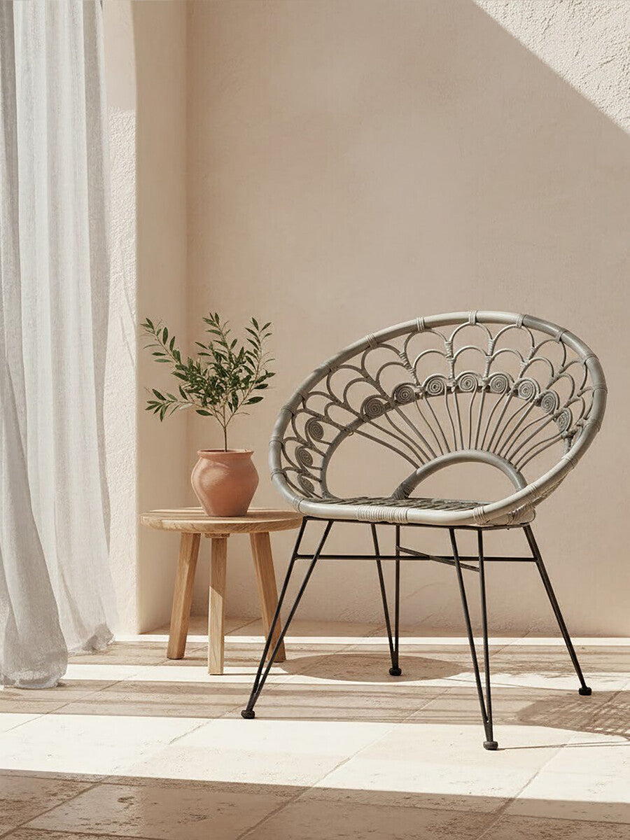 A grey natural rattan chair with a circular design, positioned next to a small wooden side table. A potted plant sits on the table, and soft sunlight filters through sheer curtains, casting gentle shadows on the floor. The background features a warm, neutral wall color