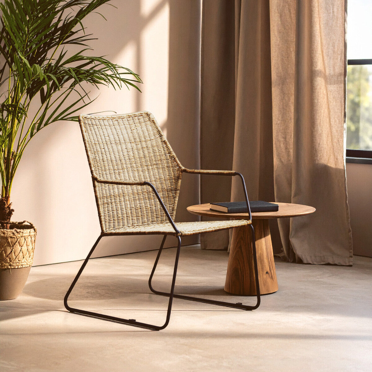 Natural rattan chair with an angled design, featuring a light-colored woven seat and backrest, supported by a sleek black metal frame. A small wooden side table is positioned next to the chair, and soft sunlight filters through sheer curtains, casting gentle shadows on the floor. A potted plant adds a touch of greenery to the scene